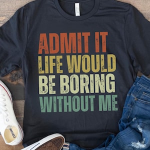 May include: A black t-shirt with the text "ADMIT IT LIFE WOULD BE BORING WITHOUT ME" in orange, yellow, and green. The shirt is paired with blue jeans and white sneakers.