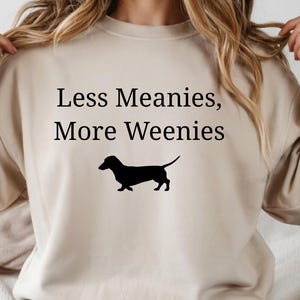 May include: Beige sweatshirt with the text "Less Meanies, More Weenies" in black, along with a black silhouette of a dachshund dog. The sweatshirt is a neutral color and has a crew neck.