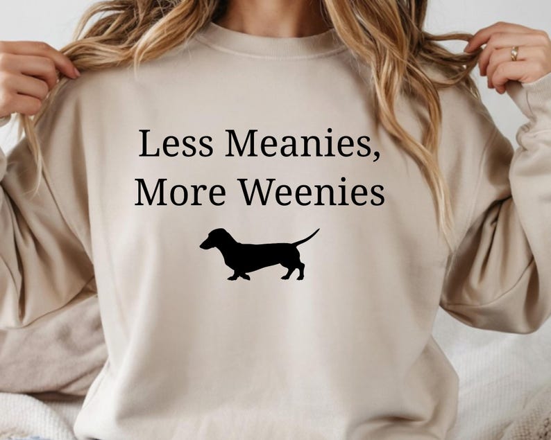 May include: Beige sweatshirt with the text "Less Meanies, More Weenies" in black, along with a black silhouette of a dachshund dog. The sweatshirt is a neutral color and has a crew neck.
