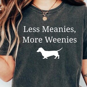 May include: Dark gray t-shirt with the text "Less Meanies, More Weenies" in white, along with a white silhouette of a dachshund dog. The shirt is a casual style.