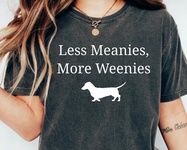 May include: Dark gray t-shirt with the text "Less Meanies, More Weenies" in white, along with a white silhouette of a dachshund dog. The shirt is a casual style.