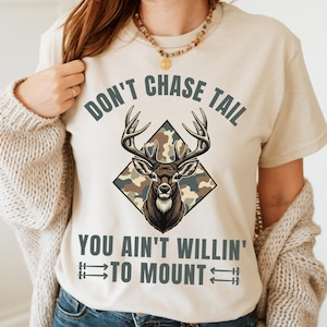 Puede incluir: Camiseta beige con un gráfico de ciervo dentro de un patrón de camuflaje en forma de diamante. El texto dice "DON'T CHASE TAIL YOU AIN'T WILLIN' TO MOUNT" en negrita. La camiseta tiene mangas cortas.