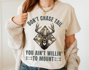 May include: Beige t-shirt with a deer graphic inside a diamond-shaped camouflage pattern. The text reads "DON'T CHASE TAIL YOU AIN'T WILLIN' TO MOUNT" in a bold, dark font. The shirt has short sleeves.