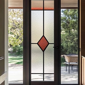 May include: A dark-framed door with a textured glass panel featuring a red diamond design. The top section has a red and tan border. The door is flanked by side panels and leads to an outdoor patio.