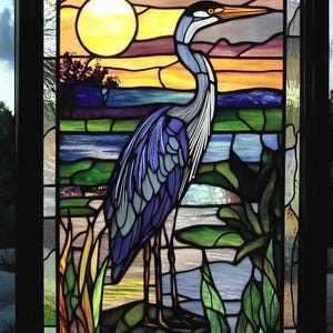May include: A stained glass window depicting a blue heron standing in a marsh at sunset. The heron has a long neck and beak, and its feathers are a mix of blue, purple, and gray. The background features a yellow sun, a blue sky, and green reeds.
