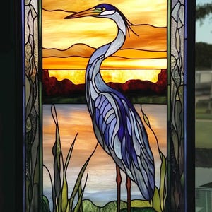 Puede incluir: Una ventana de vitral que representa una garza azul de pie en un pantano al atardecer. La garza está mirando hacia la izquierda y tiene un cuello y un pico largos. El fondo es una puesta de sol naranja y amarilla vibrante con un cielo azul arriba. El pantano es verde y marrón con juncos altos.