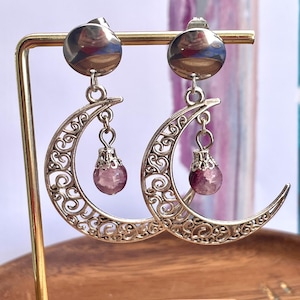 May include: A pair of silver-toned earrings featuring a circular stud top connected to a crescent moon charm with intricate filigree cutouts. A small, round, translucent purple bead hangs from the center of each crescent moon by a silver cap.
