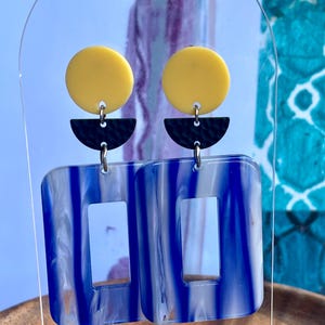 May include: A pair of dangle earrings featuring a yellow circle at the top, a black textured semi-circle in the middle, and a large rectangular pendant at the bottom with a blue and white marbled pattern and a rectangular cutout in the center.