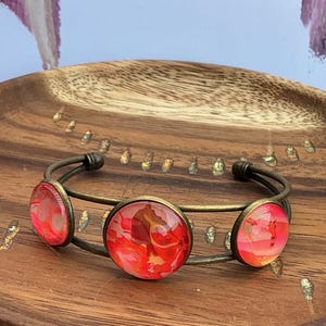 Puede incluir: Un brazalete de tono bronce con tres engastes circulares. Cada engaste contiene un cabujón liso y abombado con un patrón de remolinos abstractos y vibrantes en tonos rojo brillante, naranja y rosa suave, sobre un marco metálico.