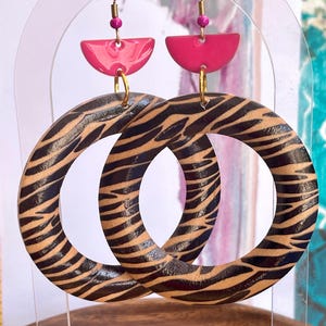 Puede incluir: Un par de pendientes colgantes con grandes aros circulares de madera con un patrón de rayas de cebra en negro y beige. Cada aro está unido a un componente de botón semicircular de color rosa brillante mediante una argolla dorada y una cuenta rosa.