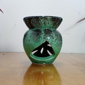 Può includere: Un bruciatore per oli essenziali in ceramica verde con disegni neri e verdi. Il bruciatore ha un corpo rotondo e un'ampia apertura nella parte superiore. È posizionato su una superficie in legno.
