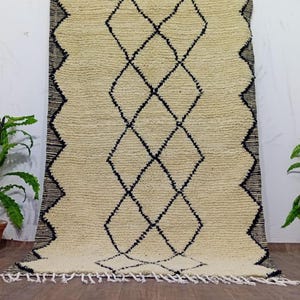 Könnte beinhalten: Cremefarbener, rechteckiger Teppich mit einem geometrischen Rautenmuster in Schwarz. Der Teppich hat eine strukturierte Oberfläche und einen Fransenbesatz am unteren Rand. Der Rand hat ein Zickzack-Design in Schwarz. Ein dekorativer Bodenbelag.