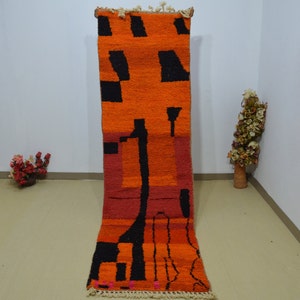 Op de afbeelding: Een lang, smal tapijt met een geometrisch patroon in zwart, oranje en rood. Het tapijt heeft aan beide uiteinden franjes.