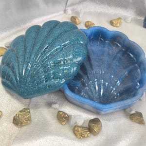 以下が含まれることがあります： 青い貝殻の形をしたキラキラ光る仕上げの小さな宝箱。箱が開いていて、内側が見える。