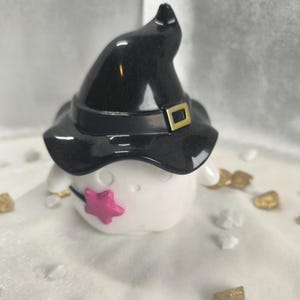 May include: A white ghost figurine wearing a black witch hat with a gold buckle. The ghost is holding a pink wand with a star on the end. The figurine is sitting on a white surface with gold and white stones.