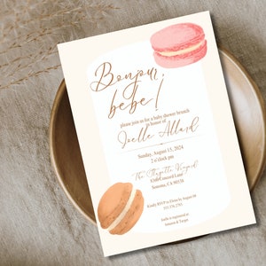 Pode incluir: Convite para chá de bebé com macarons em aquarela. O convite apresenta o texto "Bonjour, bébé!" em escrita elegante, juntamente com os detalhes do evento. O design inclui um fundo bege claro e um prato de madeira.