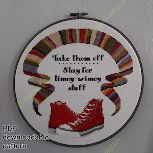 May include: A round embroidery hoop with white fabric featuring a red and blue striped scarf, two red sneakers, and the text "Take them off Stay for timey-wimey stuff".