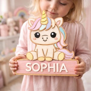 Op de afbeelding: Houten eenhoornvormige plaque met een roze basis. De eenhoorn heeft een gele hoorn en een kleurrijke manen en staart. De naam "SOPHIA" is in houten letters op de plaque geschreven. De plaque wordt door een persoon vastgehouden.