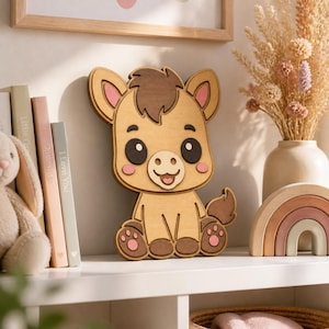 Könnte beinhalten: Ein geschichtetes Wandbild aus Holz in Form eines Cartoon-Esels mit hellbraunem Körper, dunkelbrauner Mähne und Schweif sowie zartrosa Akzenten an Ohren, Wangen und Pfoten. Der Esel hat große schwarze Augen und einen lächelnden Mund.