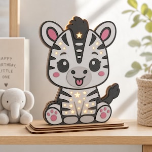 Peut inclure: Une lampe zèbre en bois superposé avec un motif rayé blanc et gris foncé, agrémenté de touches roses sur les oreilles, les joues et les pattes. La lampe présente des découpes en forme d'étoiles diffusant une lumière chaude sur un socle plat.