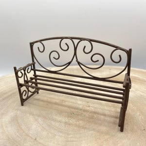Könnte beinhalten: Eine Miniatur-Metallbank in Braun mit dekorativer geschwungener Rückenlehne und Armlehnen. Die Bank hat horizontale Lamellen für die Sitzfläche und ist für ein Puppenhaus oder einen Miniaturgarten konzipiert. Die Bank ist etwa 10 cm lang.