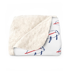 May include: White fleece throw blanket with a repeating pattern of blue pig illustrations with red lines across them.