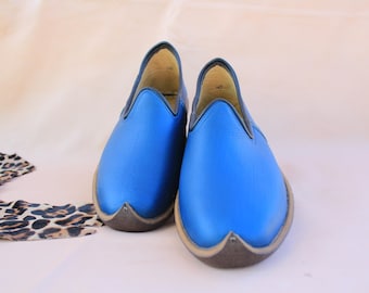 Può includere: Un paio di scarpe slip-on in pelle blu brillante con punta arrotondata e finiture scure. Le scarpe hanno un design unico e sono poste su uno sfondo neutro. Un tessuto fantasia è visibile in basso a sinistra.