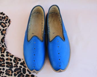 Può includere: Un paio di pantofole in pelle blu con punta a punta e un motivo di cuciture decorative. Le pantofole hanno una fodera in pelle marrone chiaro e un bordo nero attorno all'apertura.