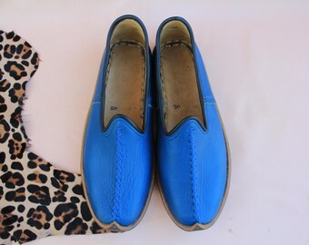 Può includere: Un paio di pantofole in pelle blu con punta a punta e una cucitura al centro. Le pantofole hanno una suola in pelle marrone.