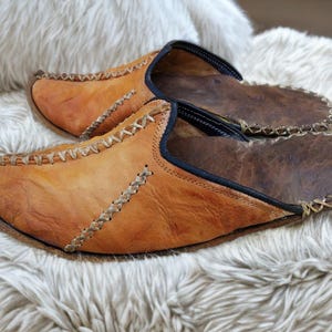 Puede incluir: Un par de zapatillas de cuero hechas a mano en tonos marrón y beige. Las zapatillas presentan costuras decorativas y un ribete azul oscuro. Este calzado de interior descansa sobre una superficie blanca y esponjosa.