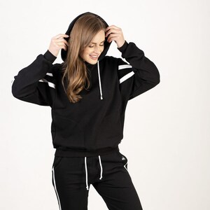 May include: A black hooded sweatshirt and matching sweatpants with white stripes down the sides. The model is wearing the set and has the hood pulled up.