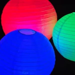 May include: Four brightly lit paper lanterns in red, blue, and green. The round lanterns have a ribbed texture, producing a soft, glowing effect against a dark backdrop. Suitable for festive decorations.