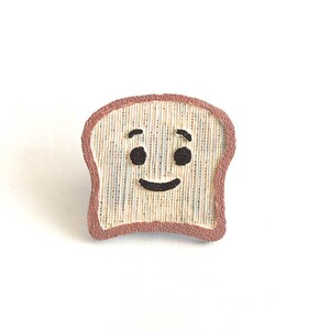 Puede incluir: Pin de rebanada de pan tostado sonriente con corteza marr&oacute;n y cara feliz. El pin es de color beige claro con l&iacute;neas verticales, ojos negros y una sonrisa. Sobre un fondo blanco.
