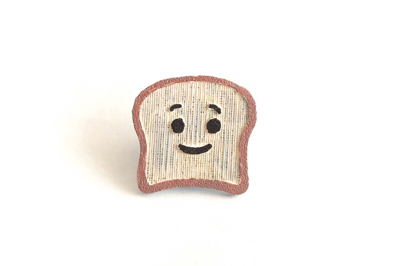 Puede incluir: Pin de rebanada de pan tostado sonriente con corteza marr&oacute;n y cara feliz. El pin es de color beige claro con l&iacute;neas verticales, ojos negros y una sonrisa. Sobre un fondo blanco.