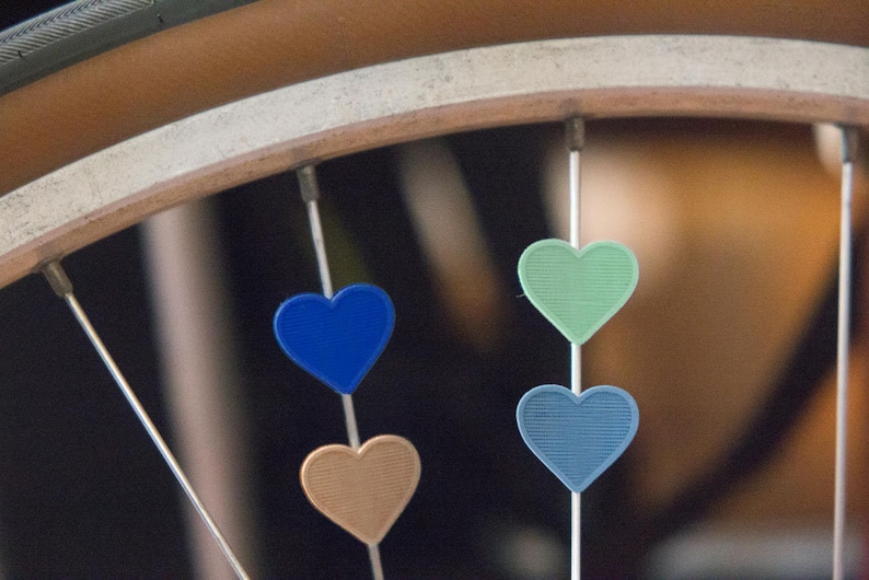 May include: A bicycle wheel with four heart-shaped charms attached to the spokes. The charms are blue, green, light blue, and brown.