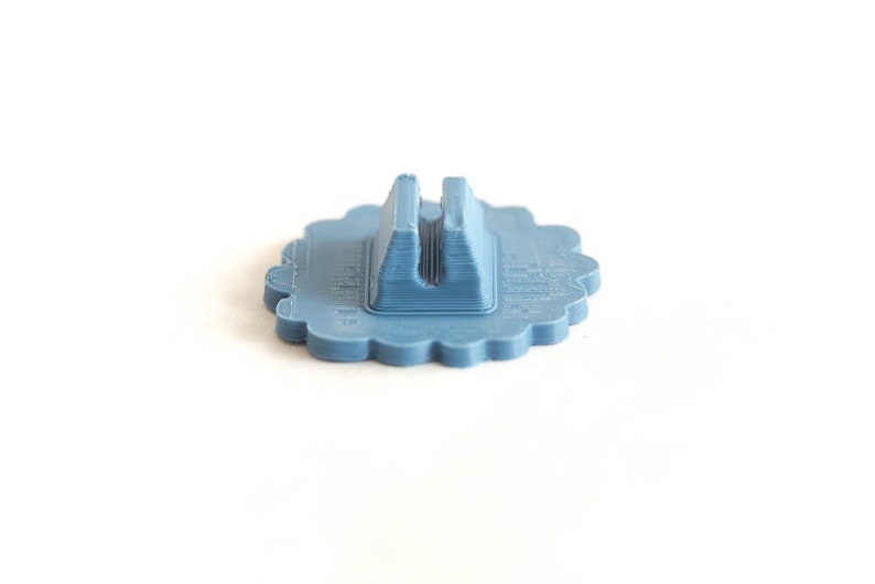 Puede incluir: Un soporte para tel&eacute;fono de color azul claro con un borde festoneado. El soporte tiene una base rectangular con una ranura central para sujetar un tel&eacute;fono en posici&oacute;n vertical. El soporte est&aacute; hecho de pl&aacute;stico y tiene un aspecto impreso en 3D.