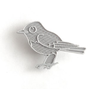 May include: A detailed, silver-colored bird pin. The pin features a stylized design of a small bird with textured lines, including the wings, tail, and body. The bird is depicted in profile, with a visible eye and beak.