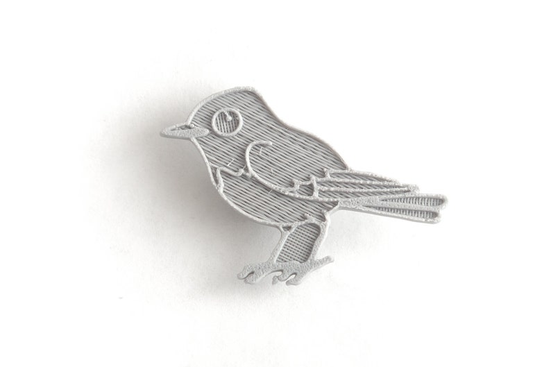 May include: A detailed, silver-colored bird pin. The pin features a stylized design of a small bird with textured lines, including the wings, tail, and body. The bird is depicted in profile, with a visible eye and beak.