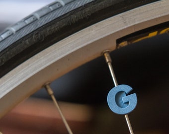 May include: Close-up of a bicycle wheel with a blue letter "G" attached to a spoke. The tire is black, and the rim is silver. The letter "G" is a light blue color.