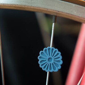 Puede incluir: Primer plano de una rueda de bicicleta con una pieza decorativa azul en forma de flor sujeta a un radio. La llanta de la rueda es plateada y el cuadro de la bicicleta es rojo. La pieza decorativa tiene un dise&ntilde;o texturizado detallado.