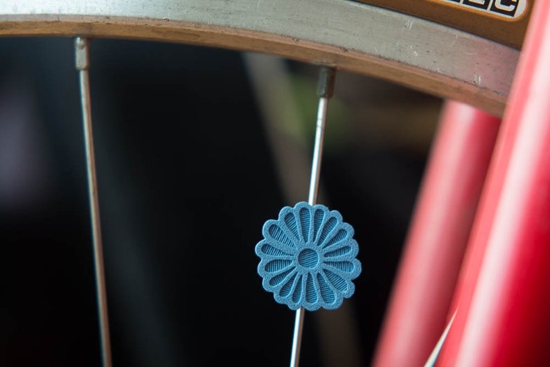 Puede incluir: Primer plano de una rueda de bicicleta con una pieza decorativa azul en forma de flor sujeta a un radio. La llanta de la rueda es plateada y el cuadro de la bicicleta es rojo. La pieza decorativa tiene un dise&ntilde;o texturizado detallado.