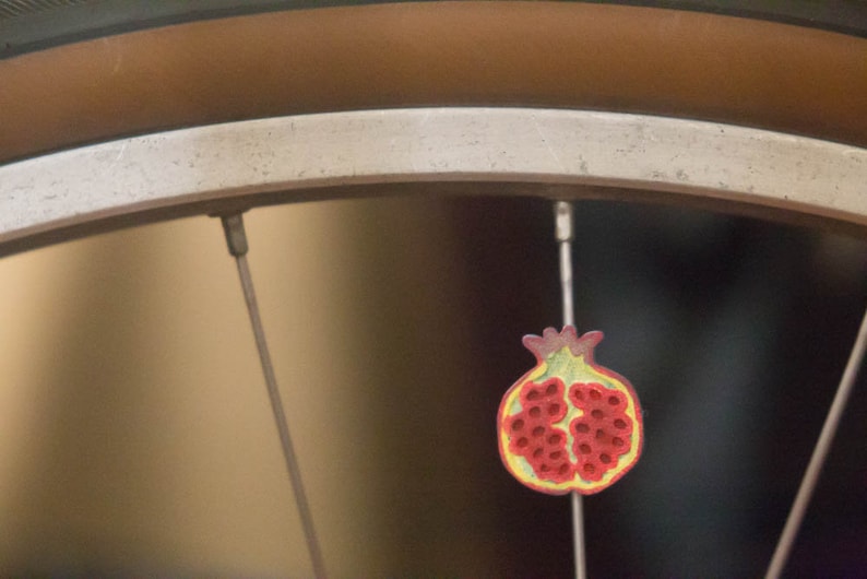Pu&ograve; includere: Un ciondolo a forma di melograno rosso e giallo appeso a un raggio di bicicletta.