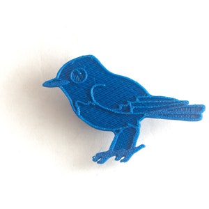 May include: A vibrant blue bird-shaped object, likely a decorative item or charm. The bird is rendered in a solid, textured material, with visible lines suggesting a 3D-printed origin. The design features a beak, eye, wings, and feet, all in a consistent shade of blue.