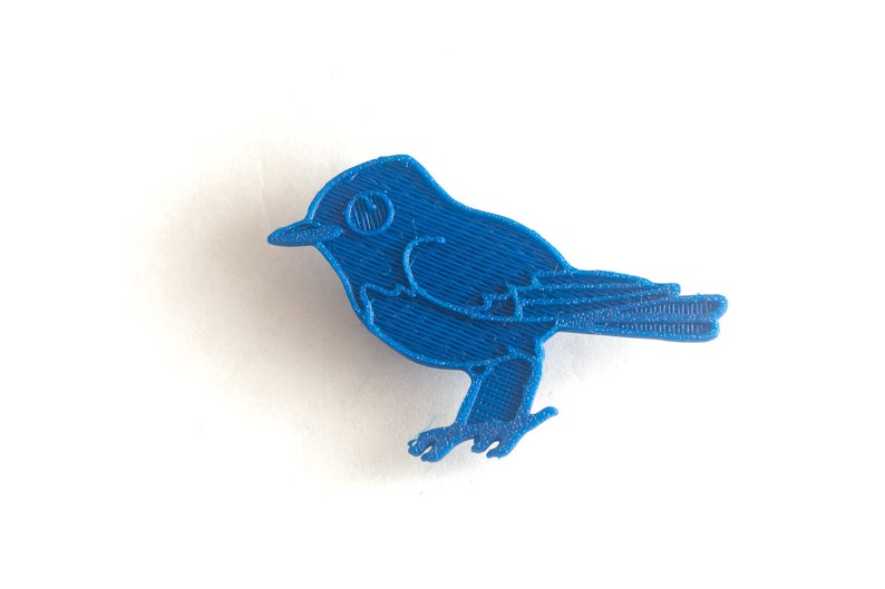 May include: A vibrant blue bird-shaped object, likely a decorative item or charm. The bird is rendered in a solid, textured material, with visible lines suggesting a 3D-printed origin. The design features a beak, eye, wings, and feet, all in a consistent shade of blue.