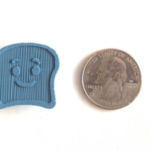 Puede incluir: Un objeto azul claro en forma de tostada sonriente con l&iacute;neas verticales, junto a una moneda de veinticinco centavos. El objeto tiene una cara de dibujos animados con dos ojos y una sonrisa curvada. La moneda es plateada.