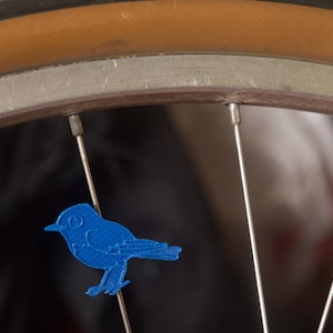 May include: A blue bird-shaped patch is attached to the spokes of a bicycle wheel. The wheel has a brown tire and silver spokes. The bird patch is a solid blue color and is facing to the right.