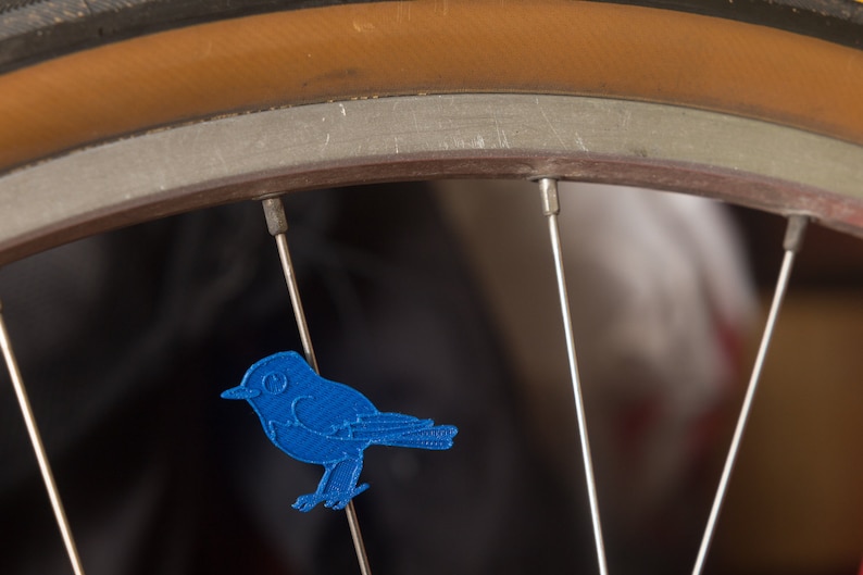 May include: A blue bird-shaped patch is attached to the spokes of a bicycle wheel. The wheel has a brown tire and silver spokes. The bird patch is a solid blue color and is facing to the right.