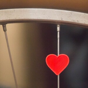 May include: A red heart-shaped charm hangs from a bicycle spoke.