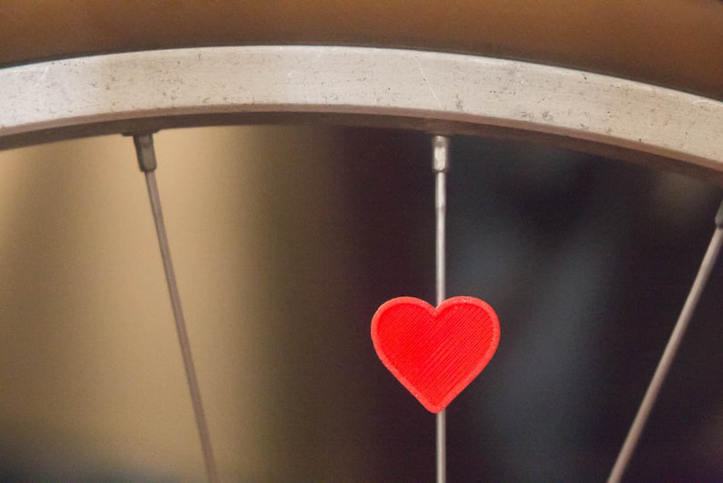 May include: A red heart-shaped charm hangs from a bicycle spoke.