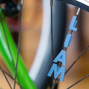 Op de afbeelding: Een close-up van een fietswiel met de naam "LIAM" gespeld in blauwe plastic letters die aan de spaken zijn bevestigd.